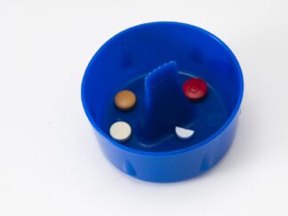 A blue plastic cup. At the centre is a flat edged spike protruding to the height of the rim, used to poke and open blister packs. Some coloured pills are lying at the bottom of the cup.