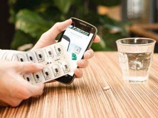 At a table, someone holds a blister pack in their hand and a smartphone in the other, with updated information on the device screen about the blister pack medications and doses removed. A cup of water and single pill sits nearby on the table.