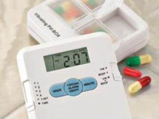 A small square white plastic device sits in front of an opened pill box, with some pills and tablets removed and laid beside it. The device has a display with the current time, sliders at either side to adjust which reminder to set or the type of reminder (e.g. vibration, sound or both). Three blue buttons sit below the display to set the hour and minute for a reminder, and one to set or turn off reminders.